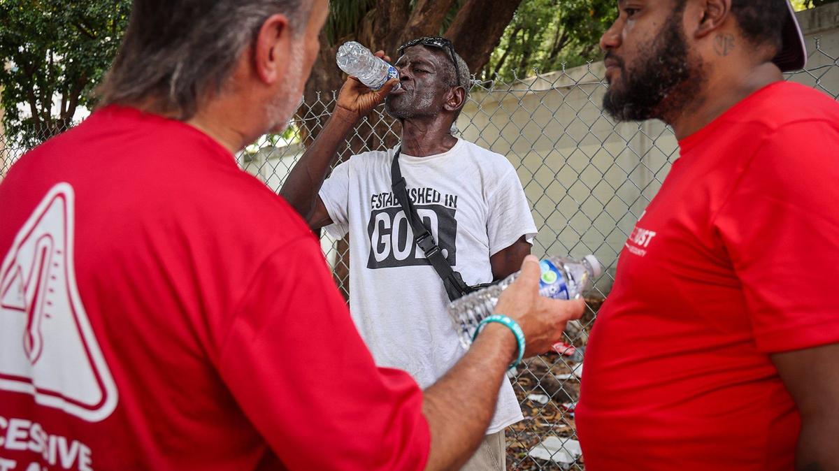 Homeless Trust Chairman Ron Book, left, and outreach worker Denny Baldonado, 40, right, stopped to speak with Juan Almeida, center. Almaida drinks a bottle of water as they survey the area and offer services to people living on the street, on Thursday, Aug. 1, 2024, in Miami, Florida.