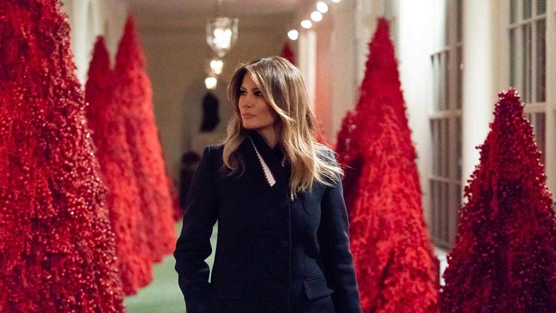 First lady Melania Trump walks through a White House corridor of red Christmas trees.