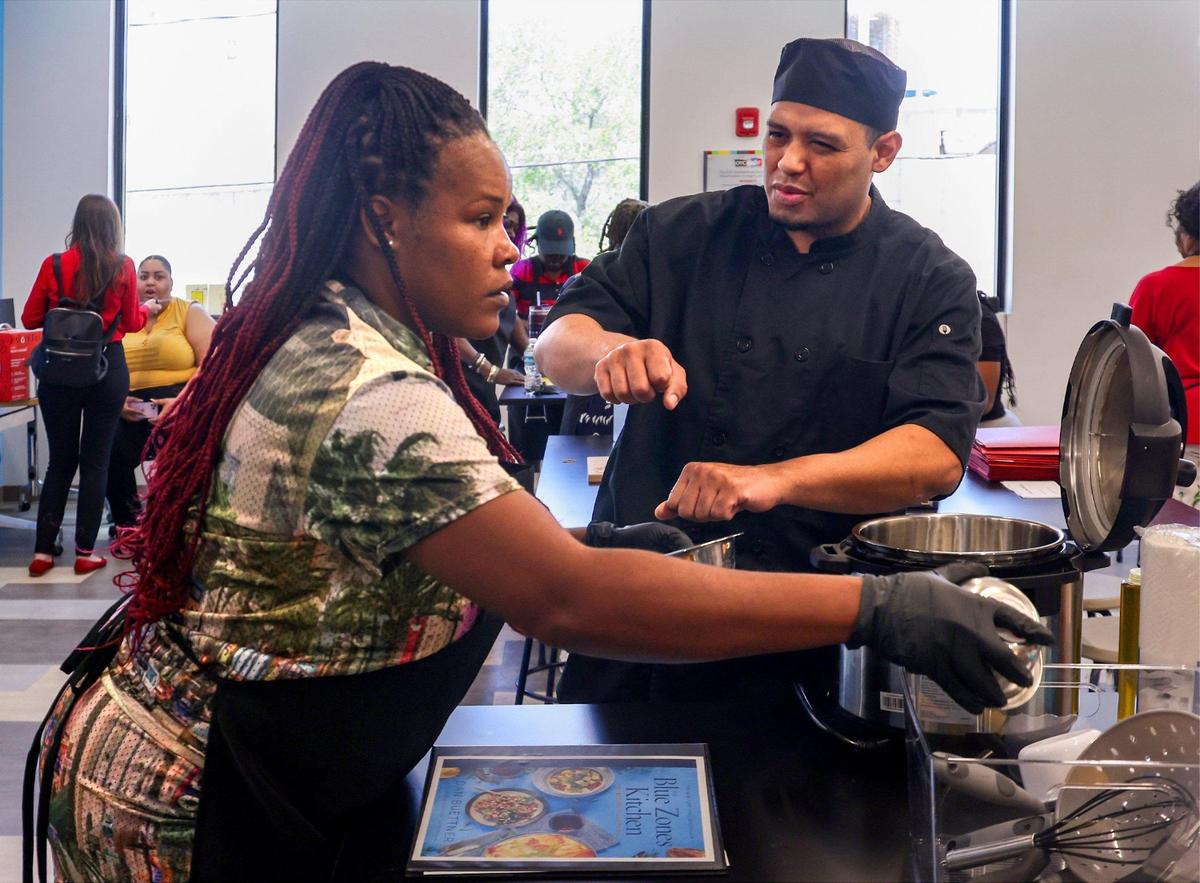 Chef Luis Montoya, 35, right, visits the “Queen of Royalty” table as Shantavous Wilcher, 40, left, requests the ingredients from members of her cooking team during a “Blue Zones” cooking class at the Overtown Youth Center on April 3, 2025, in Miami, Florida.