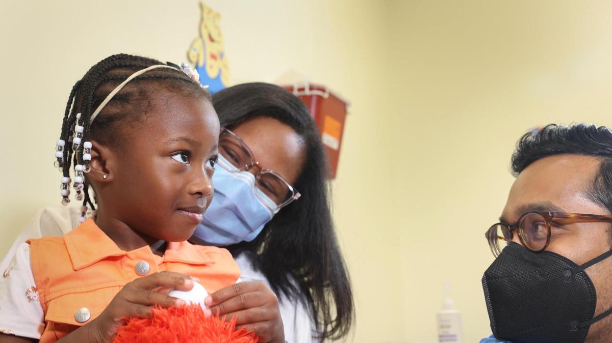 La Dra. Emy Jean-Marie sostiene a su hija, Emiola Adebayo, de 3 años, antes que el Dr. Nizar Dowla, a la derecha, le aplique la vacuna contra COVID-19 durante una campaña de vacunación para niños menores de cinco años el 28 de junio de 2022, en el Centro de Atención Médica Borinquen en Miami. Asistieron la subsecretaria de Salud, almirante Rachel Levine, y la alcaldesa de Miami-Dade, Daniella Levine Cava.