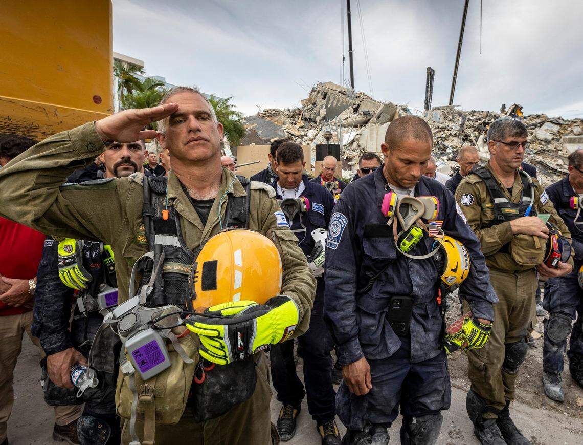 Members of the Israeli search and rescue team, left, salute in front of the rubble that once was Champlain Towers South during a prayer ceremony Wednesday night. Members of search and rescue teams and Miami-Dade Fire Rescue along with police and workers who have been working at the site of the collapse gathered for a moment of prayer and silence.