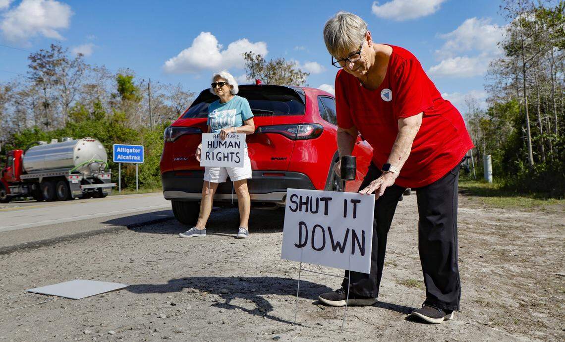 Christine Davies and Debbie Wehking, at left to right, display protest signs as vehicles drive along Tamiami Trail across the street from the front entrance of Alligator Alcatraz, on Wednesday, November 19, 2025. They are part of a  group of protesters who have been camping during the day outside of the immigration detention facility since it's opening. Alligator Alcatraz is located at Dade-Collier Training and Transition Airport inside Big Cypress National Preserve in Ochopee, Florida.