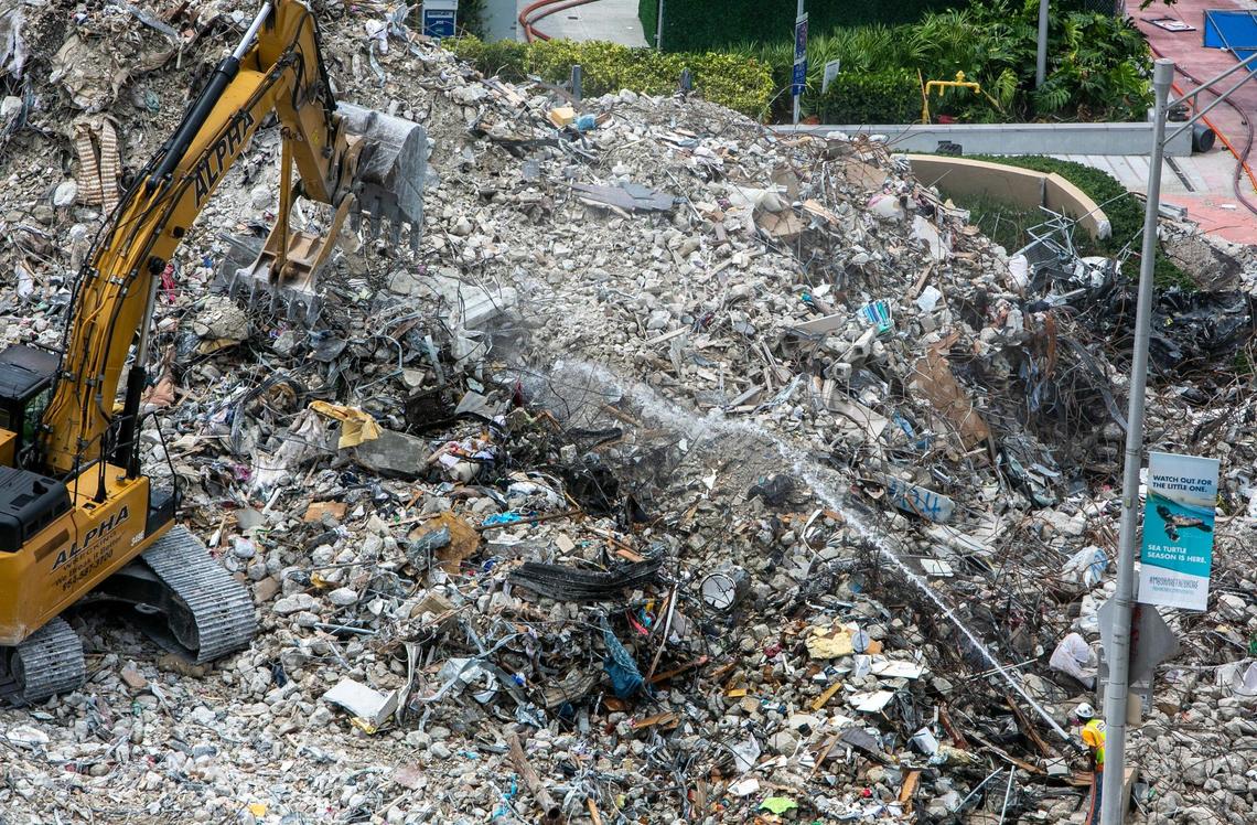 A backhoe digs through rubble on Tuesday, July 13, on the side of Champlain Towers that was imploded as another worker sprays water to keep the dust down.
