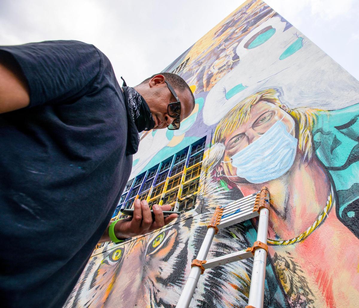 Muralist Kyle Holbrook checks a reference image on his phone for the PPE mask he just painted onto his mural of convict Joe Exotic and a tiger, from the now ubiquitous Netflix docu-series called Tiger King, at 500 NW 36th St, on Friday, April 17, 2020.