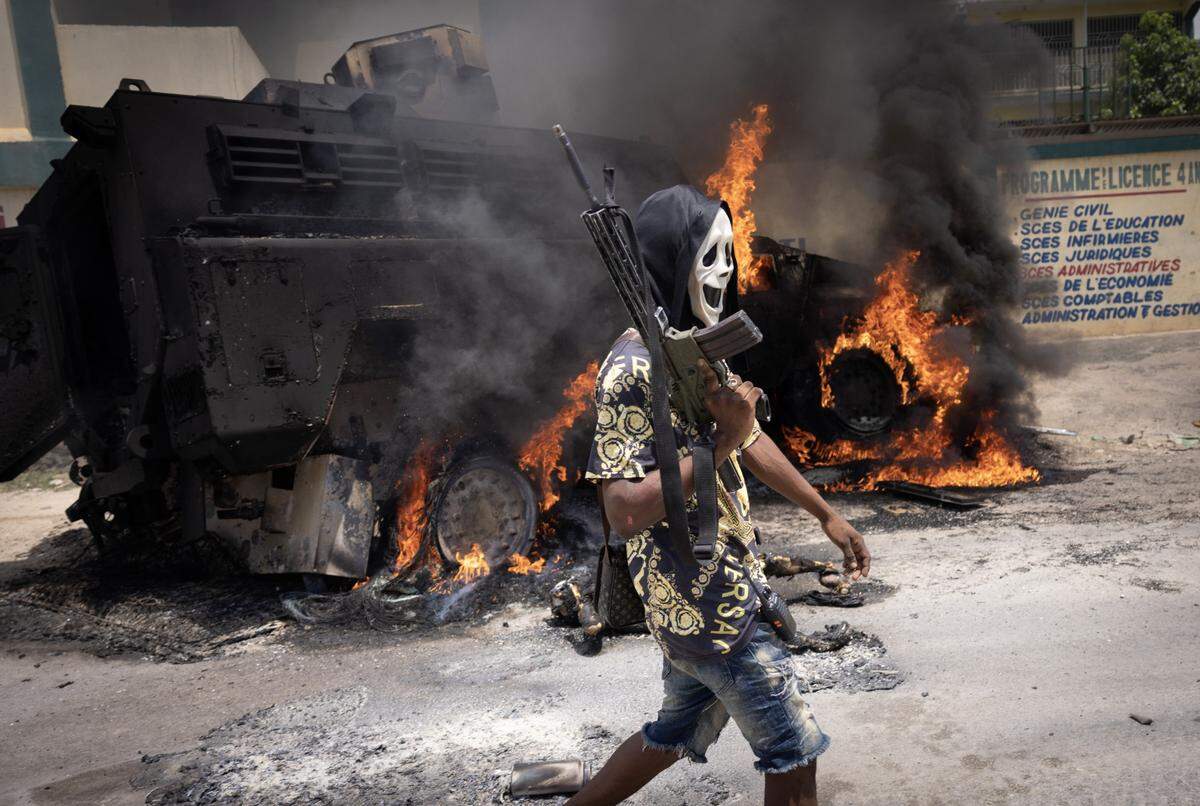 Gang members survey the husk of a police vehicle they set on fire in the town of Gressier during an attack on a police station on May 27, 2024, that resulted in the deaths of two officers, according to the gang. One member wears a vest stolen from a police officer.