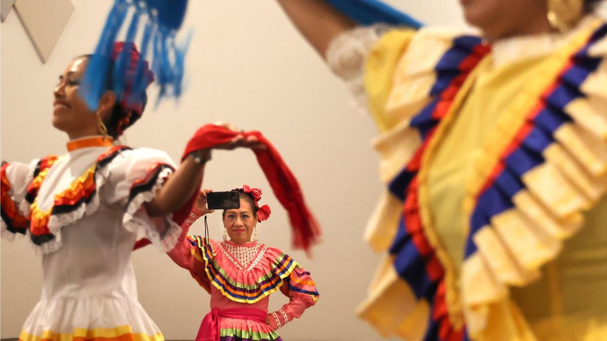 Ameyal Mexican Cultural Center’s dance company’s Livia Santiago, left, and Cecilia Ortega, right, perform on stage as Yuridia Cano, center, captures their performance on her phone. The group opened the Day of Welcome event at the Arva Parks McCabe Auditorium on Wednesday, September 18, 2024, at Miami-Dade County’s Main Library.