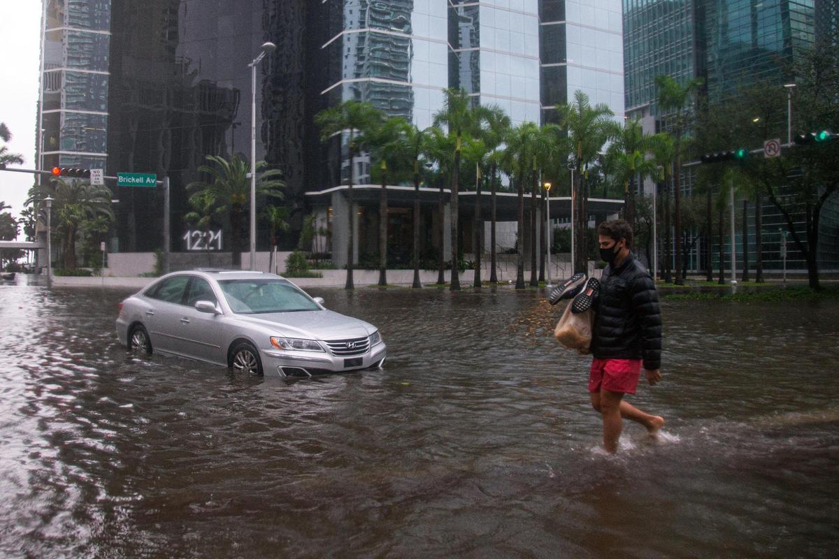Man holding a bag of groceries and a pair of shoes crosses the flooded intersection of SW 13th Street and Brickell Avenue as cars are left abandoned due to flooding brought by Tropical Storm Eta in Miami, Florida, Monday, November 9, 2020