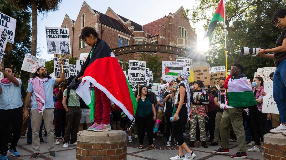 A girl wrapped in a Palestinian flag stands on a bollard as protestors advocating for the end of the siege on Gaza gather on the corner of University Avenue and 13th Street at the University of Florida in Gainesville on Oct. 25, 2023. (Gabriel Velasquez Neira/Fresh Take Florida)