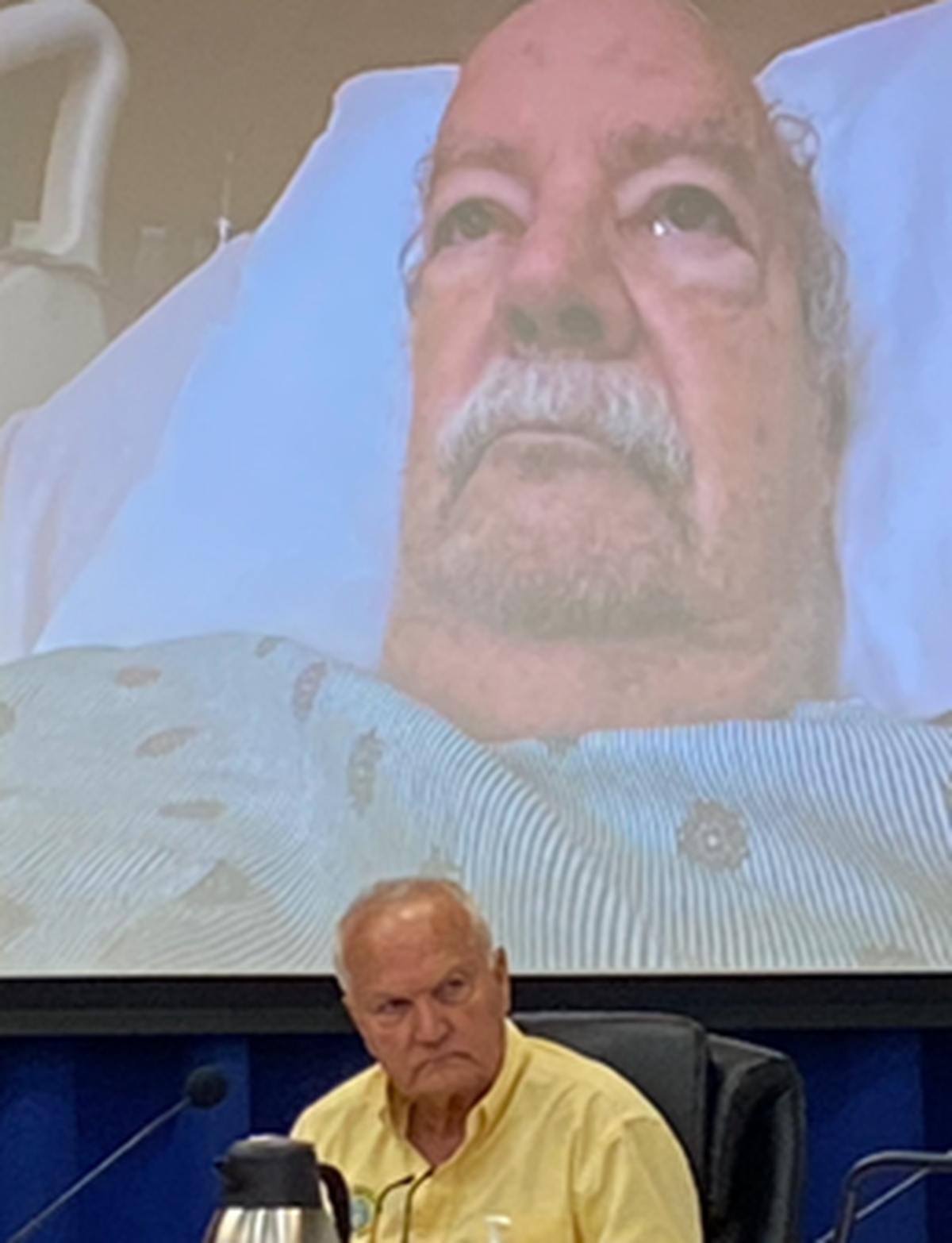 Islamorada Mayor Pete Bacheler appears on screen from his hospital bed during a Village Council meeting Thursday, Aug. 25, 2022. Sitting at the dais underneath the screen is Village Counciman Buddy Pinder. Details leading up to a vote taken during that meeting are under Monroe County State Attorney’s Office investigation.