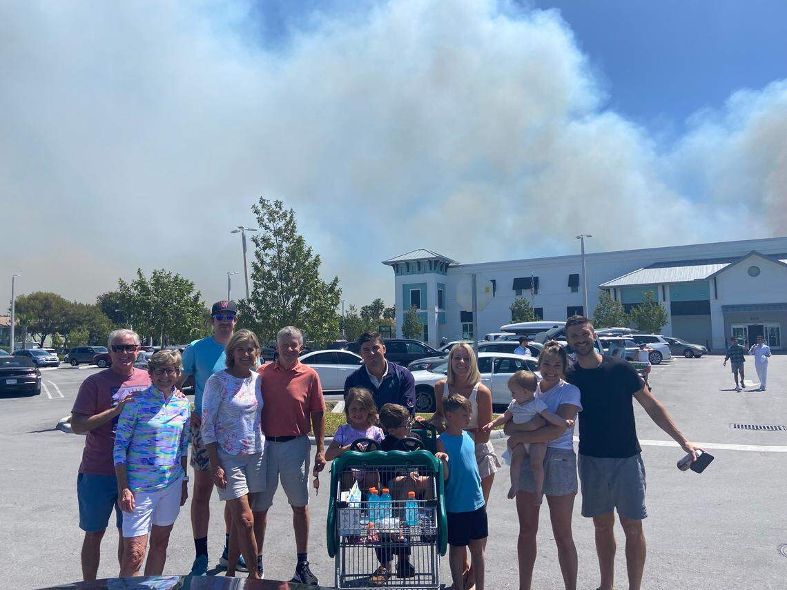 The Penellas, Follett, Self and Peirce families stand in front of the Publix at Southeast 13th Avenue in Homestead on Thursday, March 20, 2025, as large plumes of smoke from nearby brush fires loom in the background.