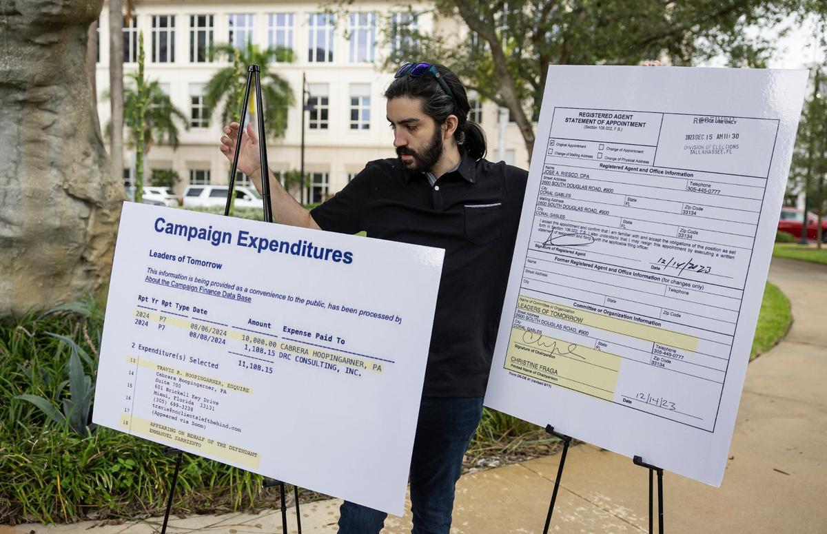Kevin Lopez holds signs as Irina Vilarino, co-owner of the Las Vegas Cuban Cuisine restaurant and a candidate for Doral City Council Seat 3, holds a press conference at Downtown Doral Park on Wednesday, Aug. 28, 2024, showing public records of Mayor Christi Fraga's involvement in a complaint challenging her candidacy as council member.