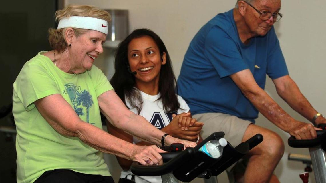 
FAMILY WORKOUT: Angela Alvarado, a health coach instructor at the UHealth Wellness Center, helps Claire Hackett, left, and her husband, Bob Hackett, cycle with a small group of people with Parkinson’s  participating in a new cycling exercise class. Studies say exercise, like cycling, proves beneficial to Parkinson’s patients. 
