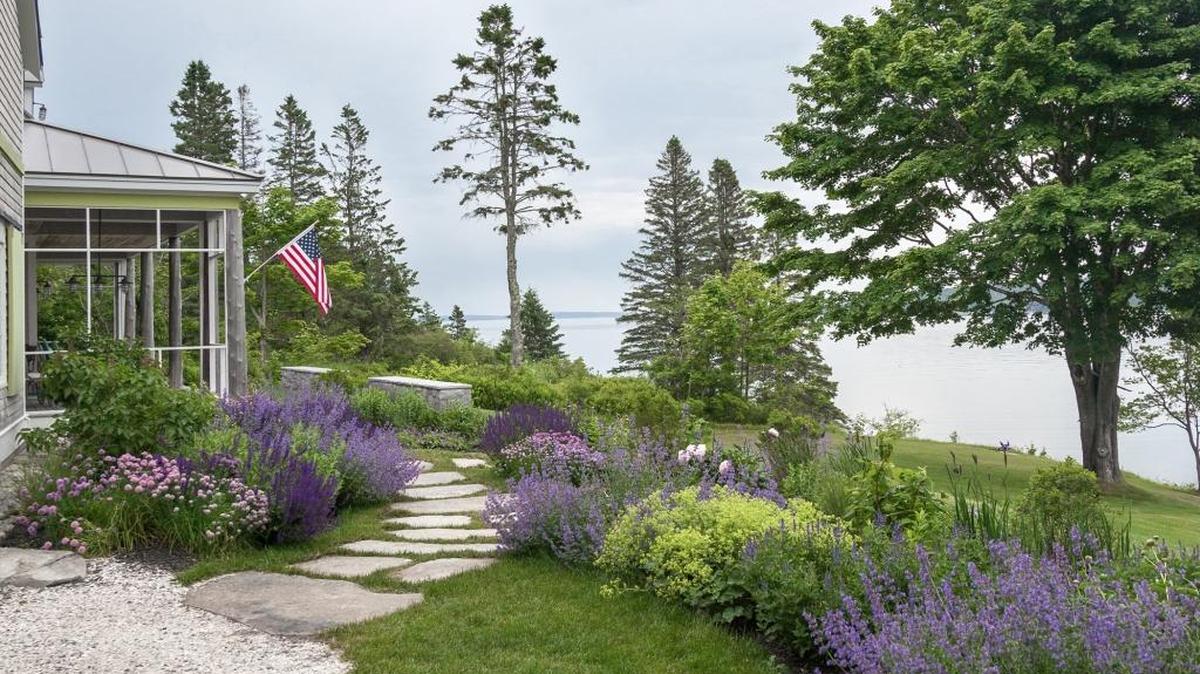 Rustic stepping-stones lead the eye through exuberant, cool-color flower beds to a low stone wall overlooking a sweeping water view. The mature trees in the background establish the natural context and lend drama to the setting.