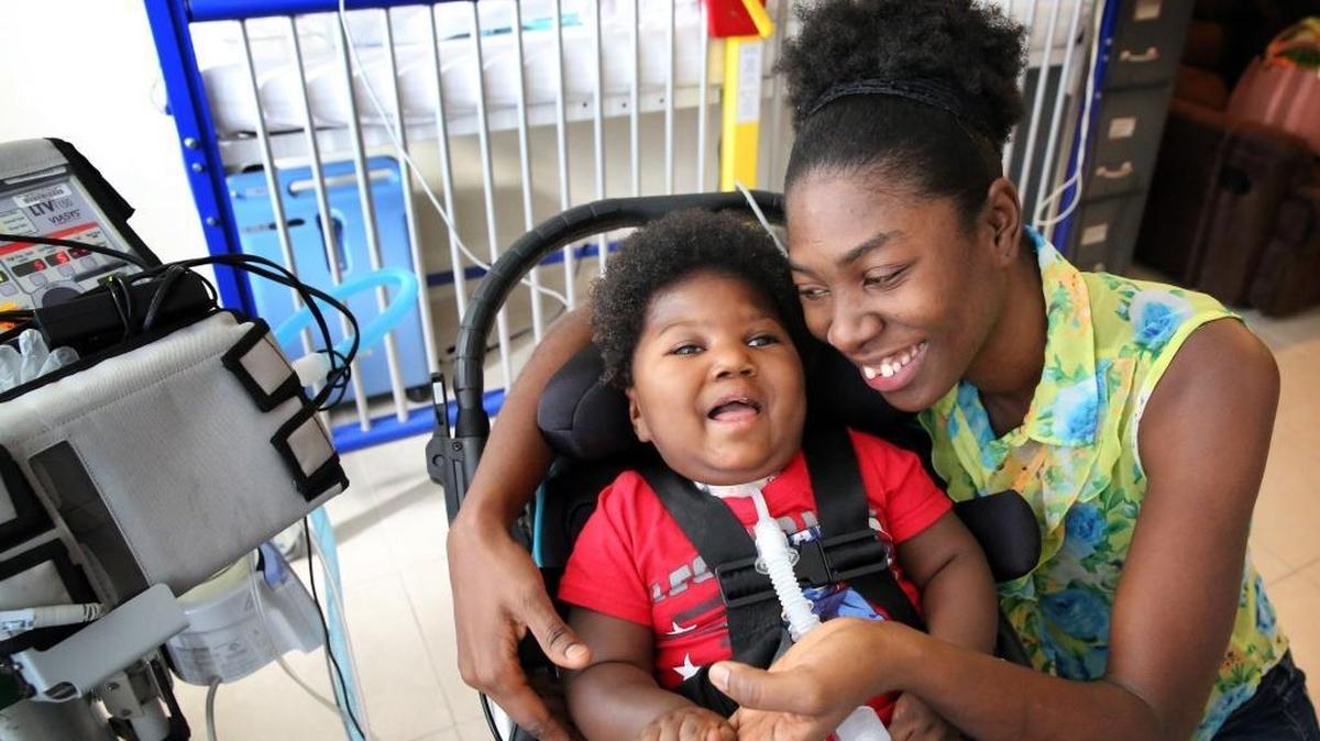 Antoinise Augustine, with her 17-month-old son, Samuel, at their North Miami studio apartment. Samuel was born prematurely with microcephaly. He remains on a respirator and is fed through a G-tube. Samuel has outgrown his infant wheelchair, so transporting him has become increasingly difficult. The family is asking for a toddler wheelchair so he can get the help that might make it possible for him to one day attend school.