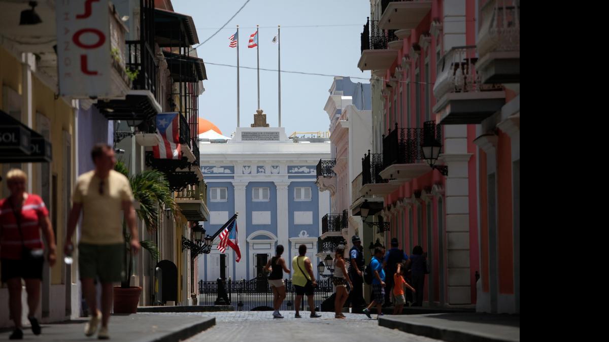 
People walk on the street in front of the governor’s mansion on the background in Old San Juan, Puerto Rico, Monday, June 29, 2015. International economists released a critical report on Puerto Rico's economy Monday on the heels of the governor's warning that the island can't pay its $72 billion public debt.
