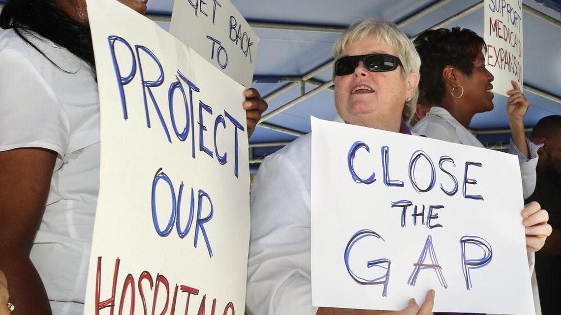 Longtime Jackson Health System registered nurses Lisa Ward, left, and Barbara Scollon, center, hold hand-lettered signs during a press conference and rally supporting Medicaid expansion in May 2015. Florida’s Legislature, meeting this month in Tallahassee, is not likely to debate the question of Medicaid expansion this year, observers say.
