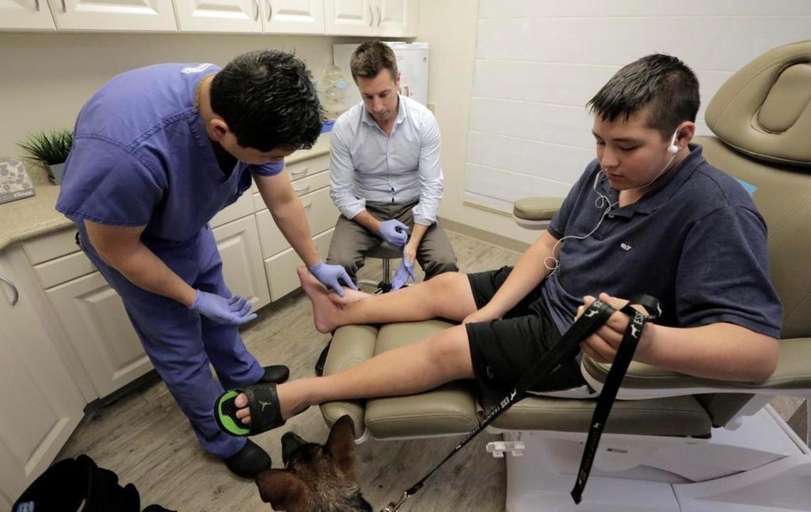 Doctors Michael Cheung (left) and Christopher Low (center), of CRS Plastic Surgery in Fort Lauderdale, examine Kyle Laman’s AR-15 bullet wound during an April 27 visit. The doctors worked to repair the damage to Kyle’s right ankle by patching the wound with a section of skin and muscle taken from the teen’s thigh. Two months after the surgery, the wound has healed, they said.