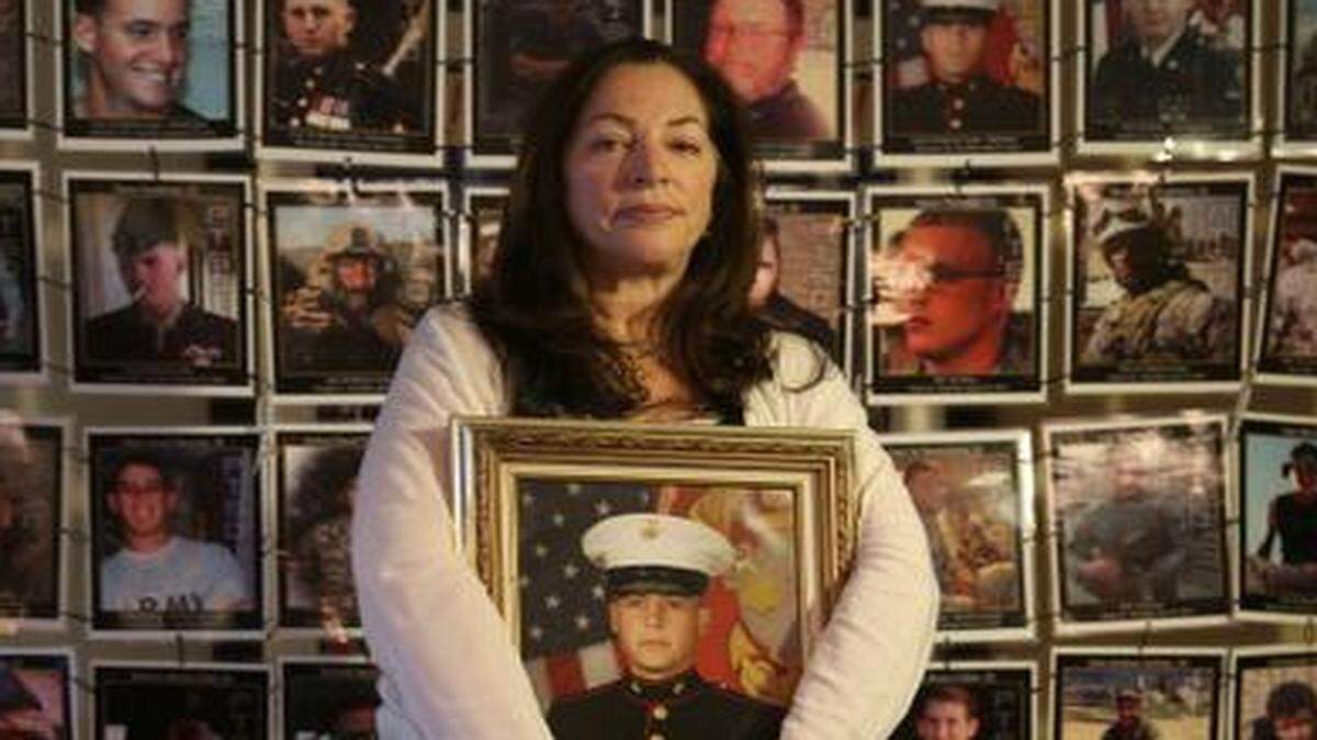 
Janine Lutz, the mother of a U.S. Marine who committed suicide last year, is planning to sue the VA. She blames the VA for prescribing her son, Janos Lutz, drugs that he told doctors made him feel suicidal. She is surrounded by photos of veterans who have taken their lives. She is holding a Marine portrait of her son.
