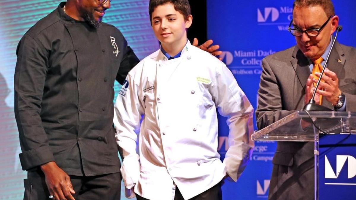 
Chef Richard Ingraham with Seth Munday, an 18-year-old battling Crohn’s Disease. The two stood on stage after Ingraham helped Munday put on a culinary jacket at Miami Dade College’s Miami Culinary Institute at the college’s Wolfson campus. Munday’s wish to meet his culinary idol, Ingraham, came true during the Chef Coat Ceremony welcoming the latest class of culinary students. Dr. Jose Vicente, Wolfson campus president, applauds.
