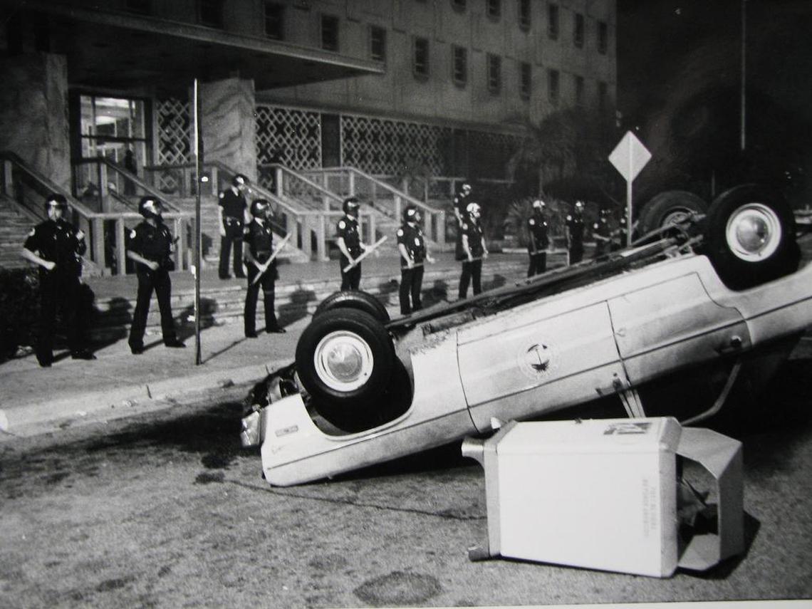A police car sits toppled as riot police stand guard.