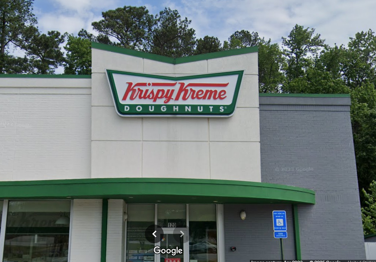 A close-up, street-level view of a Krispy Kreme Doughnuts shop. The building is white and gray brick with a green stripe at the top and bottom, and a green and red Krispy Kreme logo sign is centered on the facade.