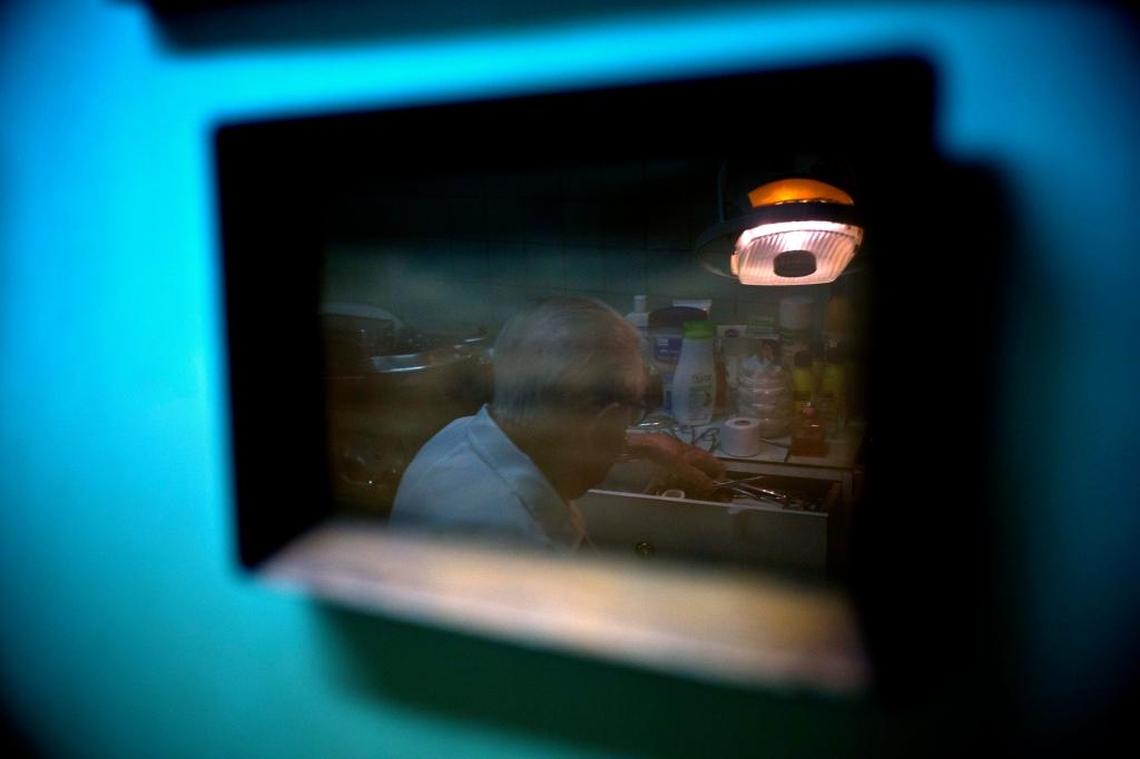 In this June. 8, 2017 photo, podiatrist Serafin Barca is seen reflected on a picture frame as he attends a patient in his clinic in Havana, Cuba. For a dollar, Serafin Barca will spend a half hour cutting the corns off a senior citizen's foot, or an hour treating a twisted ankle.