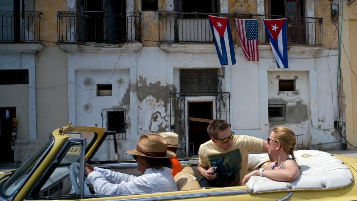 Havana visitors ride in a classic American car past a balcony decorated with Cuban and U.S. flags in Havana, Cuba. After a series of hikes in hotel prices, rates are starting to come back down to earth.