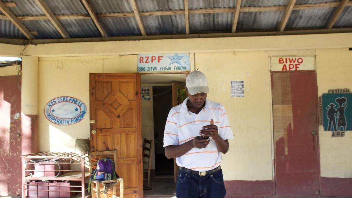 Enel Beaulière stands in front of his radio studio in Fondwa, Haiti, looking at his phone for someone to call about troubles with launching Radio Zetwal’s broadcast. Behind him, the open door to the studio.