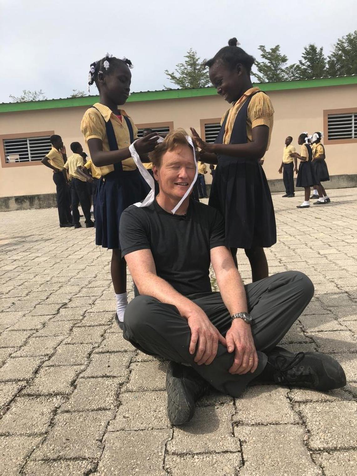 During a visit to a school in Haiti — Ecole NouvelleZoranje — comedian Conan O’Brien gets some grooming from students who decided to decorate his hair with white ribbons. O’Brien recently visited Haiti to film his “Conan Without Borders” show.