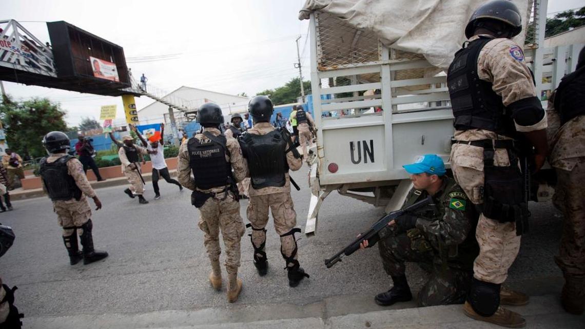 A UN Peacekeeper takes cover behind national police officers while demonstrators throw rocks, during a protest against the country's electoral council to mark the 25th anniversary of first democratic election in 1990, in Port-au-Prince, Haiti, Wednesday, Dec. 16, 2015. The demonstrators started throwing rocks after UN Peacekeepers from Brazil fired teargas into the crowd.