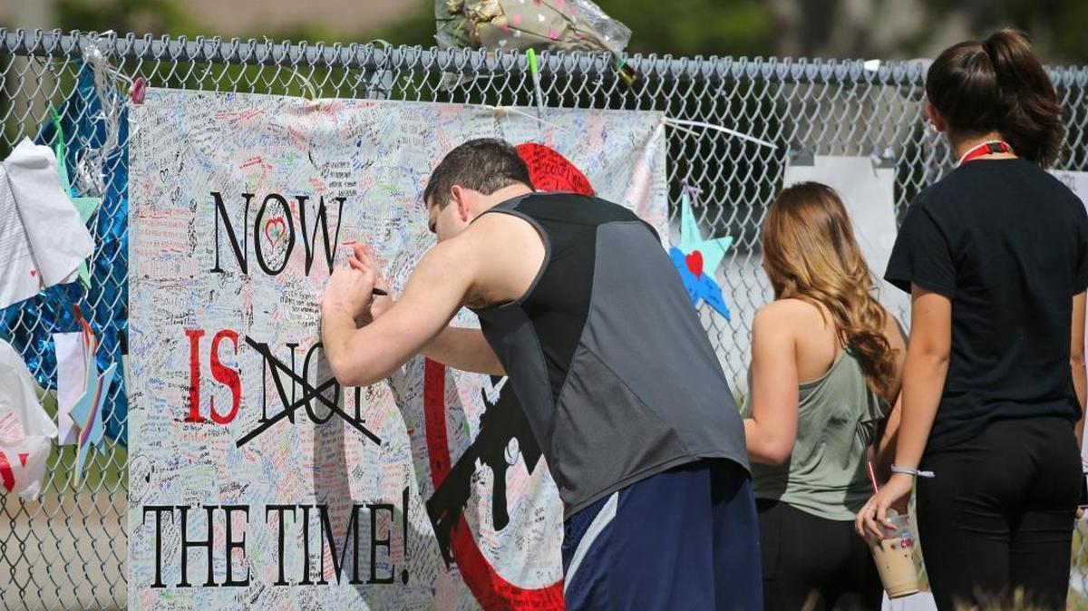 A man signs a ''No more guns'' banner as mourners pay tribute at a memorial for the victims of the shooting at Marjory Stoneman Douglas High School on Sunday, February 25, 2018, during an open house as parents and students returned to the school for the first time since 17 people were killed in a mass shooting at the school in Parkland on February 14, 2018.