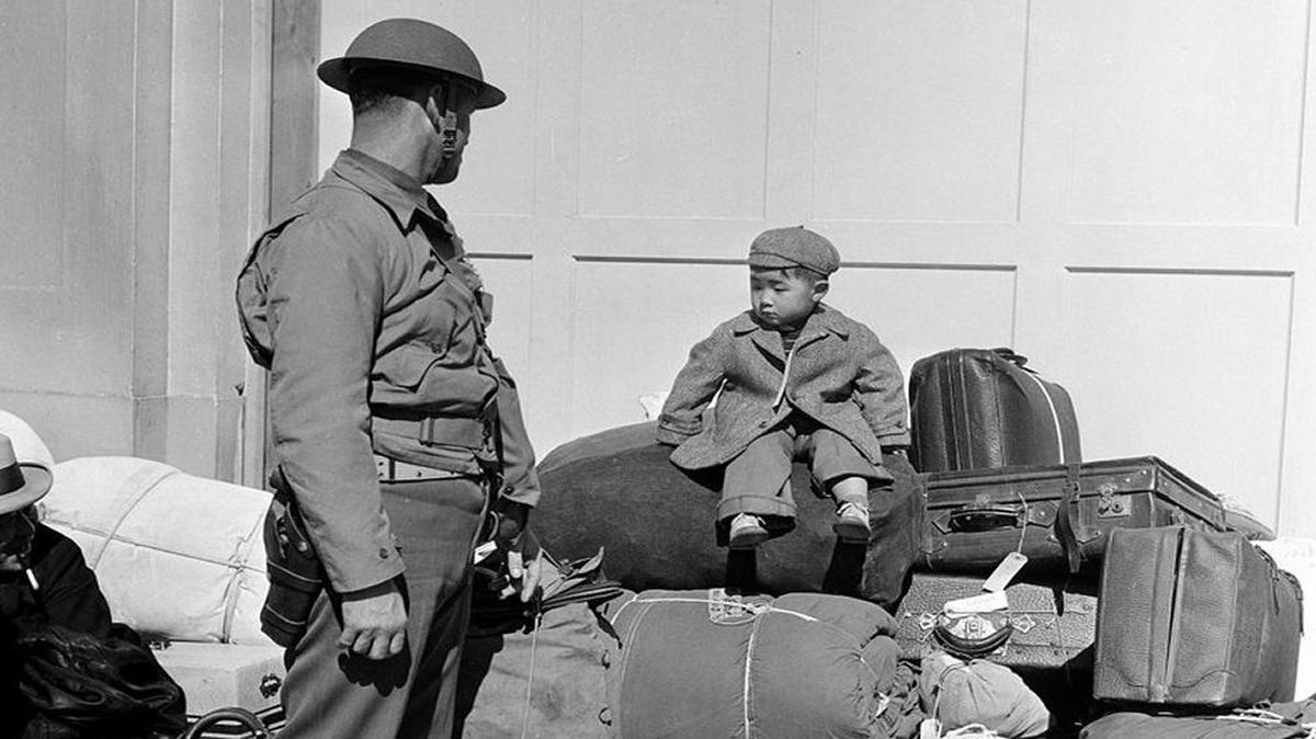 In 1942, a military officer watches as a Japanese-American boy sits on a pile of luggage, waiting for his parents, who are being processed for internment in Santa Anita, California.