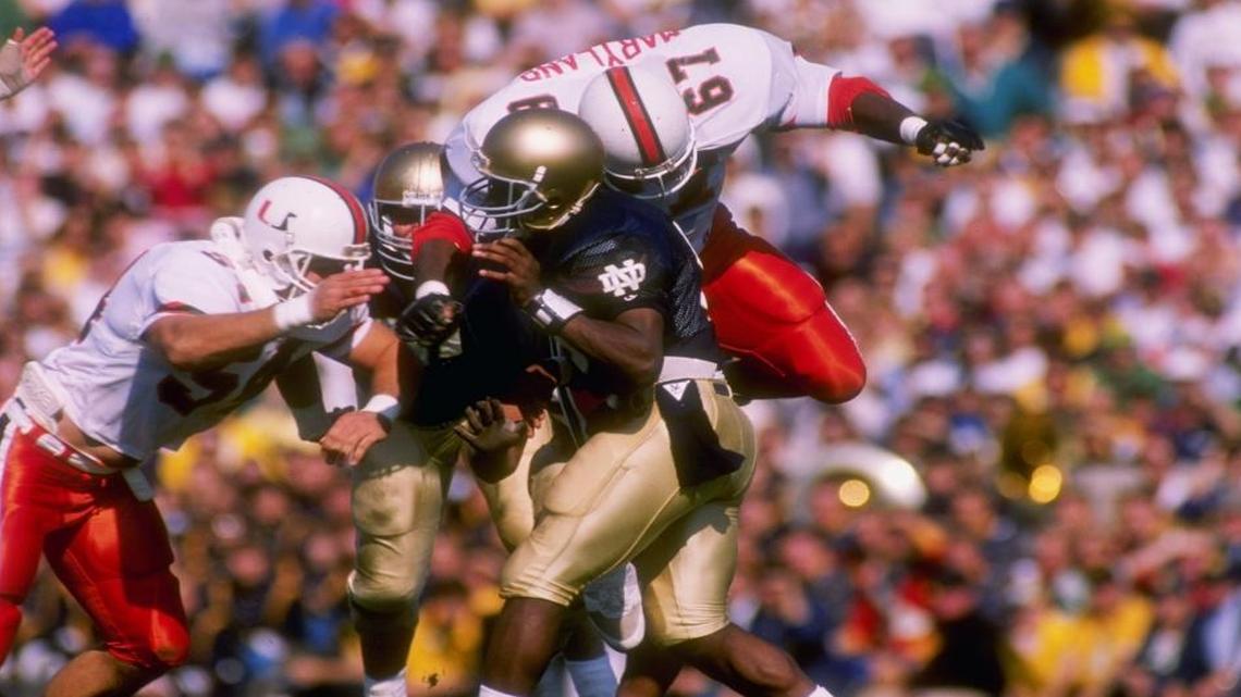 Defensive tackle Russell Maryland, 67, and defensive end Bill Hawkins, 54, of the Miami Hurricanes hit quarterback Tony Rice of the Notre Dame Fighting Irish during a game against the Fighting Irish at Notre Dame Stadium in South Bend, Indiana in 1988. Notre Dame won the game 31-30.